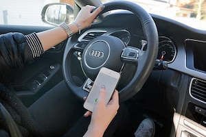 Benutzung eines Mobiltelefons am Steuer eines PKW. Eine Hand am Lenkrad, die andere hält ein Handy, Smartphone, Mobiltelefon.