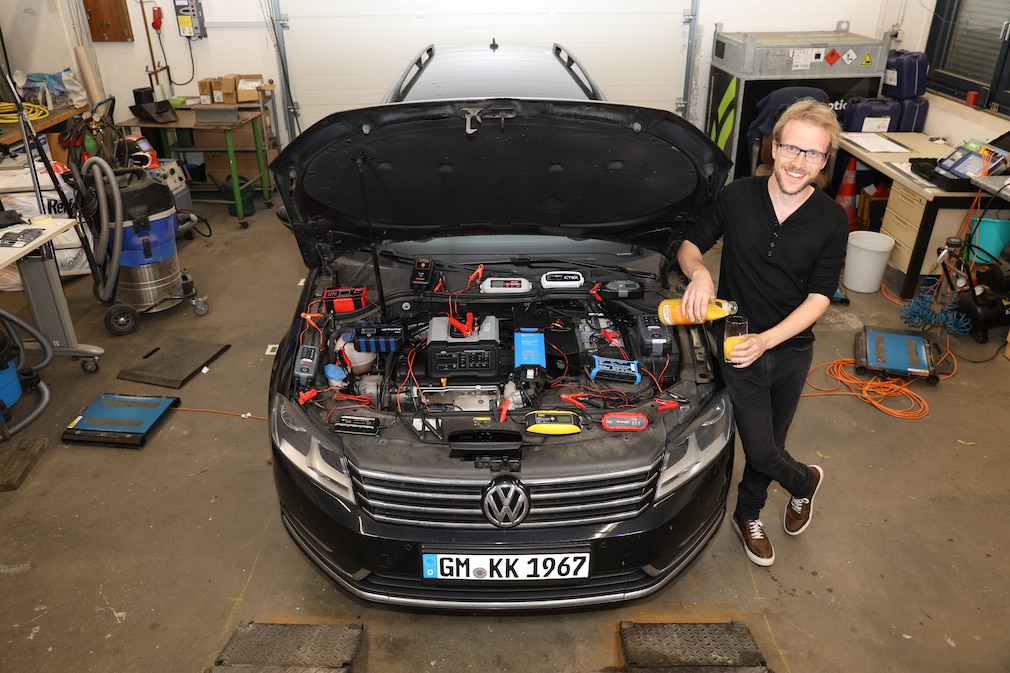 Person lehnt am vorderen Kotflügel eines Autos und schenkt sich ein Glas Orangensaft ein. Die Motorhaube ist geöffnet und im Motorraum verteilt sind alle getesteten Ladegeräte.